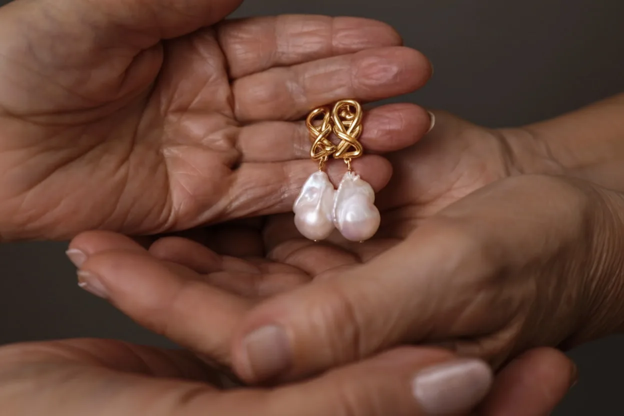 Pendientes de autor MUSUBI con diseño entrelazado y perlas barrocas, reposando entre las manos de una pareja mayor como símbolo de una joya que trasciende generaciones.